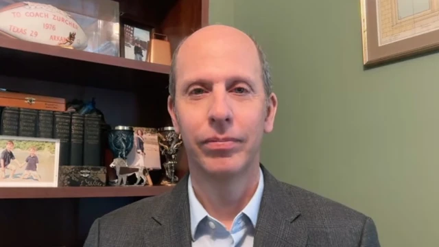 A man in a pale blue shirt and grey jacket looks directly at the camera.