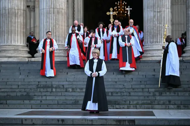 Mulllally dressed in robes at the bottom of St.Paul's steps with religious clergymembers in red and black robes descending the stairs behind her, in January in London.