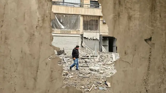A man is photographed through a hole in the wall as he walks along a street strewn with building debris at the site of an overnight Israeli airstrike