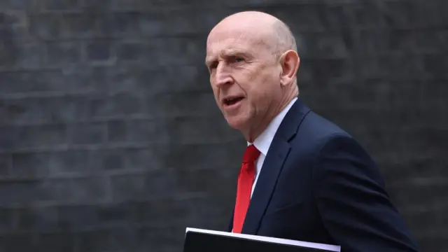 John Healey is seen walking while holding a folder and wearing a red tie
