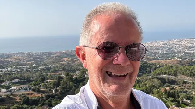 A man wearing sunglasses and a white shirt smiles with a coastal town behind him, with trees and the sea visible among houses.