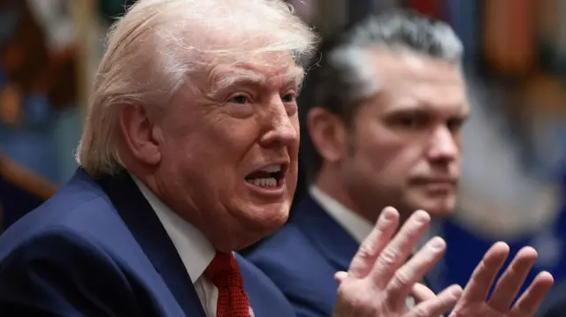 Donald Trump speaking at a press conference and gesticulates with his hands while Defense Secretary Pete Hegseth is blurred in the background next to him