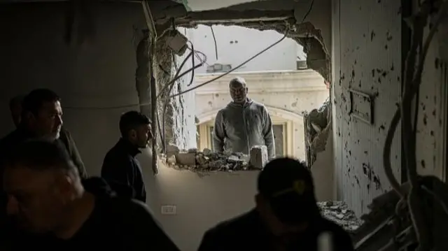 A man stares through a round hall in the wall as people look on from the other side. Several wires have become visible and there are several loose bricks