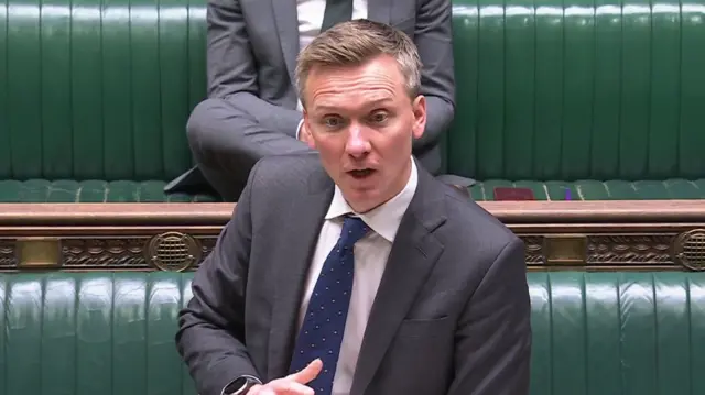 Pensions minister Torsten Bell mid-speech as he delivers speech in the House of Commons. He's standing up, leaning his left arm of a wooden lectern while wearing a dark grey suit. Behind him are the green leather benches of the Commons, a colleague in a grey suit sitting down behind him