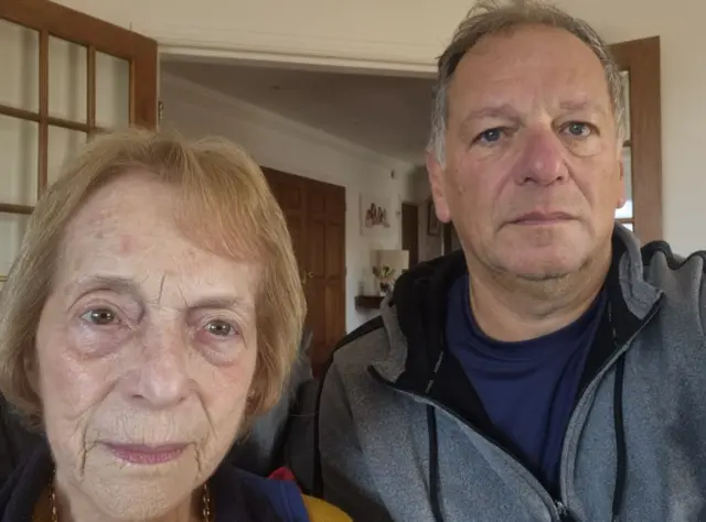 A selfie taken by Phil Fraser of himself and his mother, Ruth. Phil has short grey hair and wears a grey zip-up jumper over a navy t-shirt. Ruth has short red hair and wears a top with a blue collar. They are stood in front of a set of internal double doors.