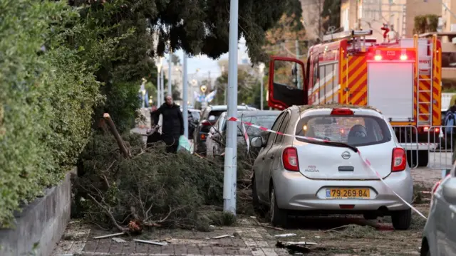 Tree branches lie on a pavement with emergency tape and an emergency vehicle nearby.