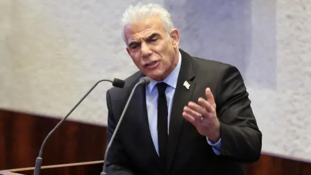 Israeli opposition leader Yair Lapid speaking into two microphones at a podium and raising his left hand