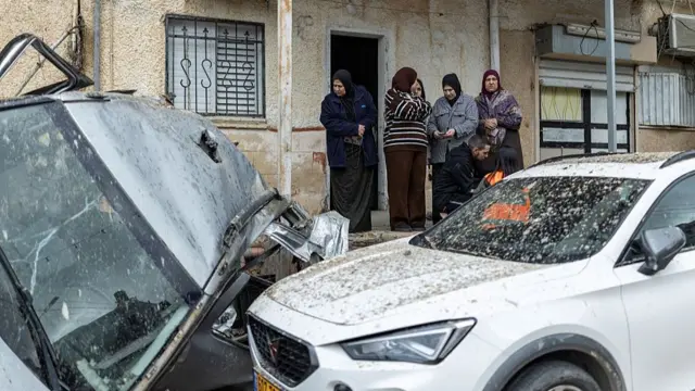 A group of people stand on the porch of a house and look over two severely damaged cars