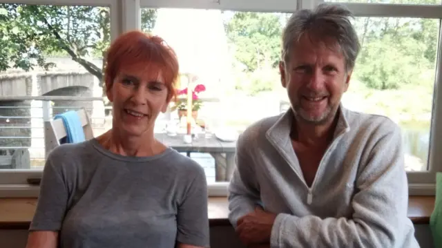 Rhona Edwards and her husband Alex, smiling for a photo in front of a large window. Rhona has short red hair and wears a grey t-shirt. Alex has short grey hair and wears a grey quarter-zip fleece. Through the window behind them, it is a sunny day and a row of trees can be seen.