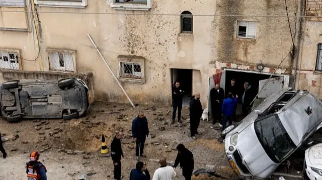 A group of men inspect where a strike hit a street in Kfar Kasem. Rubble from the strike can be seen sprayed against a wall and two cars have been flipped over