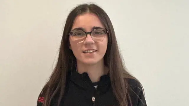 Lucy Johnston looks directly at the camera with a plain white background behind her. She is a white woman with long brunette hair and is wearing glasses. She is wearing a zip up black jacket.