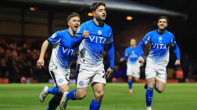 Stockport's Josh Stokes, in County's home blue kit, celebrates a goal to make it 1-0 against Northampton on 17 March by running away with his hand raised. His team-mates can be seen running after him in the background.