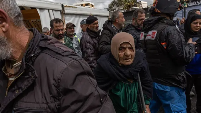 An elderly woman in a brown headscarf is standing in line to receive food from tents.