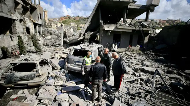 Four men standing in a pile of rubble and destroyed buildings with a green hillside and cloudy sky in background