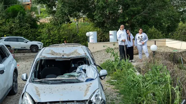 Civilians in Beit Shemesh view a damaged car near a crater left by a missile