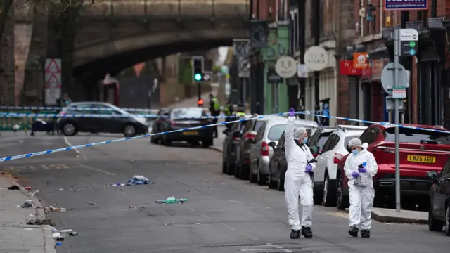 Two forensic officers dressed in white walk under a blue and white police cordon.