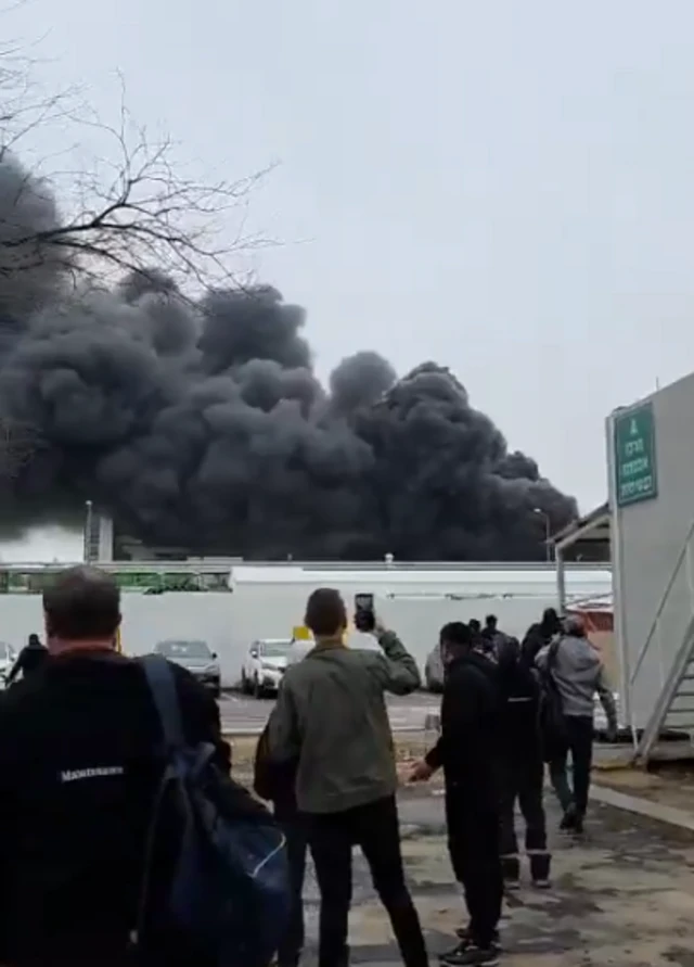 Large plumes of smoke billow upwards as a group of people watch on from a distance. One person is filming the smoke with their phone.