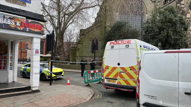 Two uniformed police officers stood between a patrol car and police van next to a blue and white cordon.