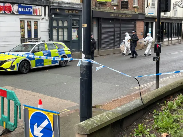 Two people in white forensic suits carrying two plastic bags each with two uniformed police officers walking close by.