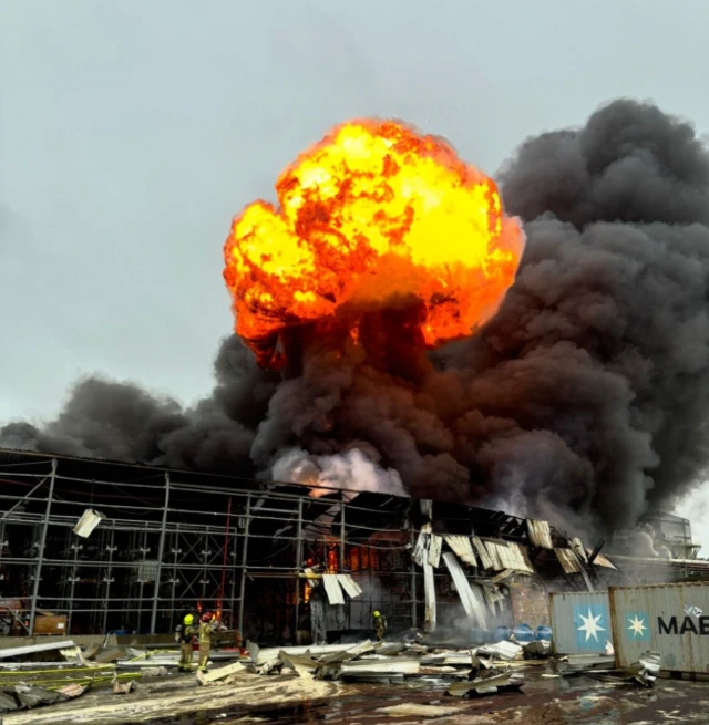 A large ball of fire and heavy black smoke erupts from the site of the fire, covering most of the sky