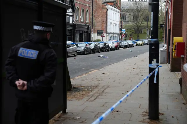 A police officer stood next to a blue and white cordon surrounding a road with some debris on it.