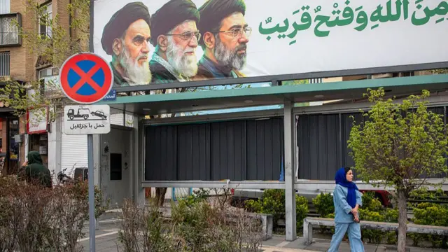 A woman stands walks by a poster featuring the current and former Supreme Leaders on March 28, 2026 in Tehran, Iran.