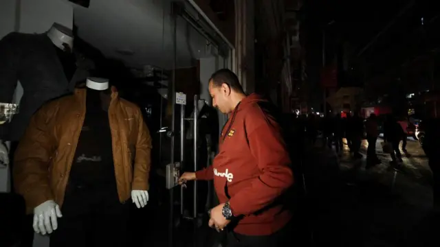 A man wearing a red hoodie locks the door to his clothes store. In the background several silhouettes can be seen in the darkness
