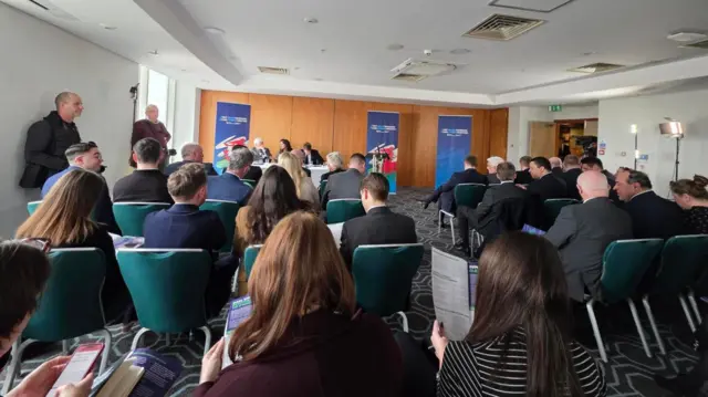 a picture from the back of the room showing the gathered reporters in front of the podium