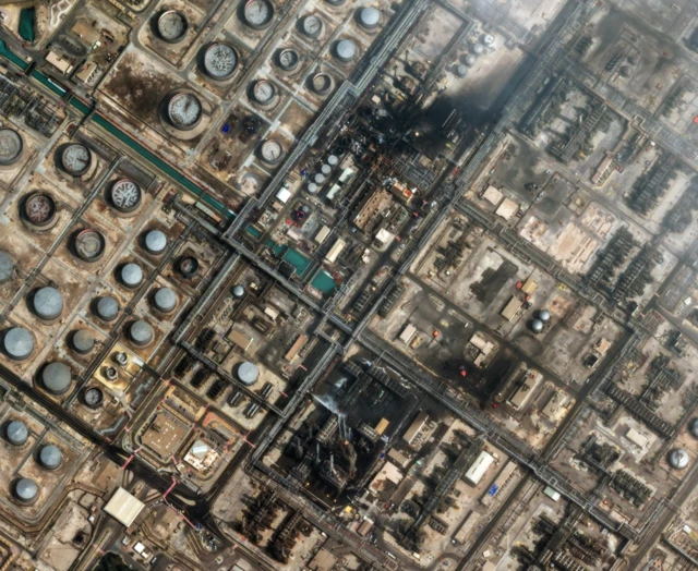 A satellite photograph shows two blackened areas of oil-refinery infrastructure that was hit by a drone strike. There appear to be streams of white water or fire-extinguishing liquid being sprayed on one of the blackened areas, which sits amid a wider complex of pipes, tanks, sheds and paved roads.