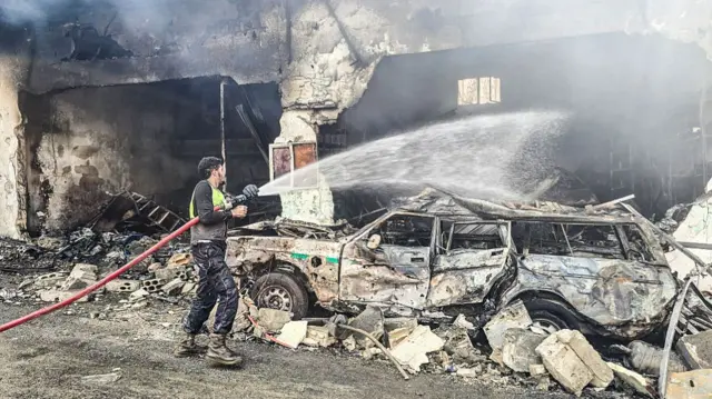 a worker hoses a burned car and collapsed building with water in Hanouiyeh