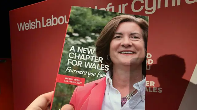 A hand can be seen holding the election manifesto, which has a photo of Eluned Morgan smiling and includes the words 'A new chapter for Wales' and 'fairness you feel'. It is pictured against a Welsh Labour red backdrop.