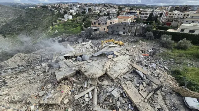 a hillside covered in collapsed buildings and rubble, with village and hills int he background