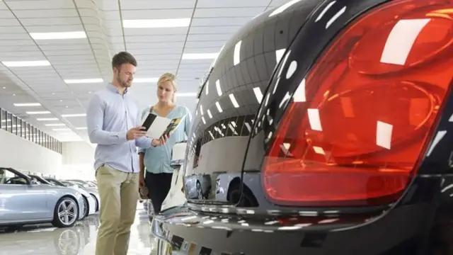 A man and women in the middle distance of an indoor car dealership stand looking a brochure, with the boot of a black care in the foreground.