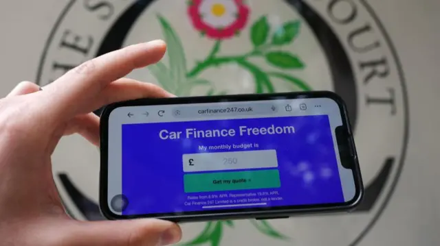 Left hand holding am iPhone looking at the Car Finance Freedom website. The logo of the Supre Court is painted on the wall in the background.