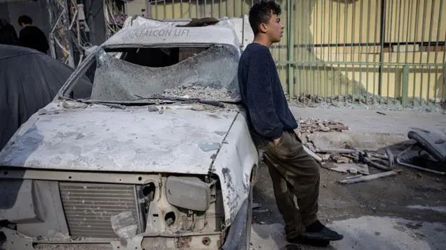 a boy leans against a damaged car surrounded by rubble on a street