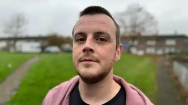Man in black t-shirt and pink hoodie stood in front of large patch of grass and row of houses
