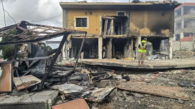 a severely damaged house in front of a large pile of rubble and a collapsed structure, with worker standing nearby