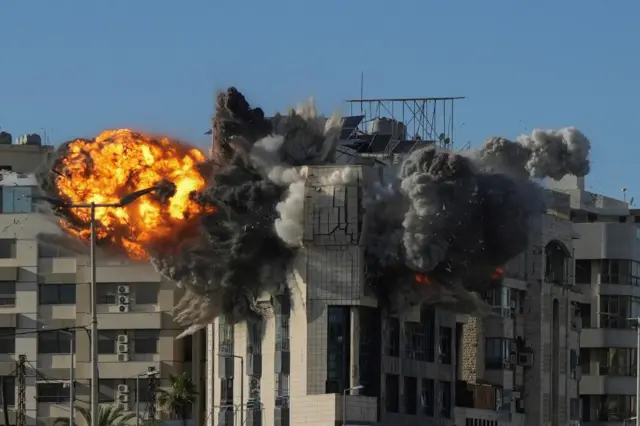 Flames and smoke burst from the top floor of a multi-level residential building as it is hit in an airstrike. The picture appears to show the moment of impact, with the building's brickwork beginning to crumple outwards.