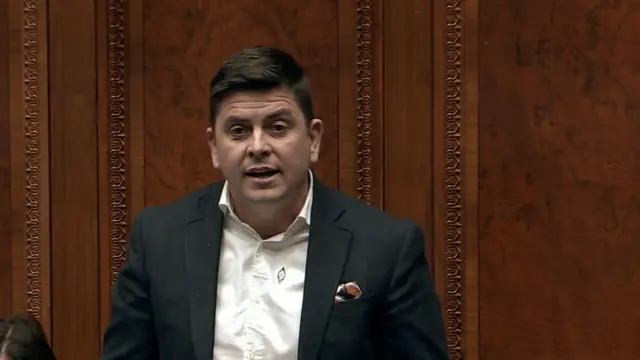 Danny Baker, with short black hair, wearing a white shirt and a dark grey suit.