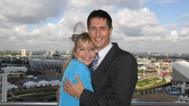 Chris and Carol embrace while posing on a balcony overlooking a large stadium and cityscape, with clouds and blue sky in the background
