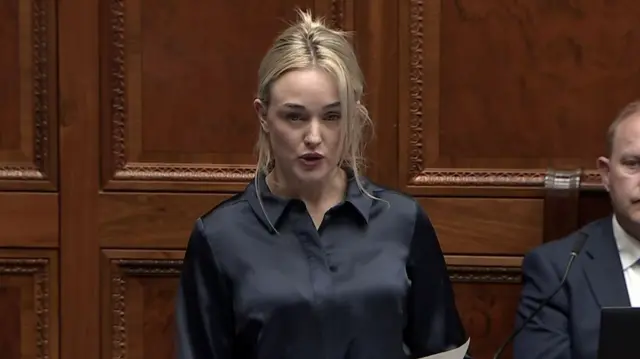 A woman in a black shirt with blonde hair addresses the assembly, holding a sheet of paper.