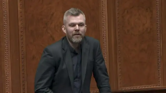 Gerry Carroll, wearing a black shirt and a black suit, standing in the Assembly chamber.