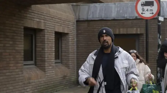 Ashley Warren is seen in front of a brick wall. He is wearing a white jacket and black hat.