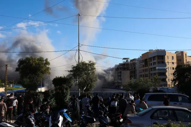 Crowds gather on a residential street, watching a smoke cloud up the road from a strike on a building. They are standing amid vehicles that have come to a standstill.