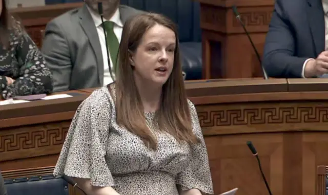 Nuala Mcallister, with long brown hair, wearing a white and black dress. She is standing in the Assembly chamber.