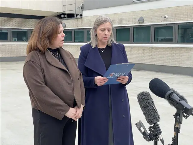 Two women dressed in coats, holding a clipboard standing in front of three microphones reading a statement
