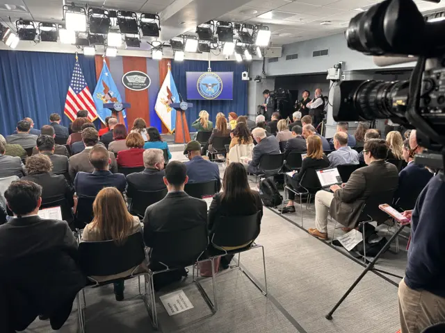 A busy room. Photo is taken from the back and shows rows of chairs filled with people all facing away from the camera towards a small stage with two podiums on it