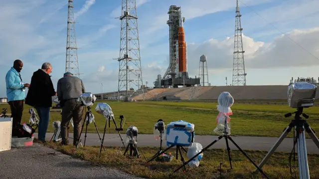 In the foreground, cameras are set up outside facing a rocket ship in the background. Three men stand on the left near one of the cameras.