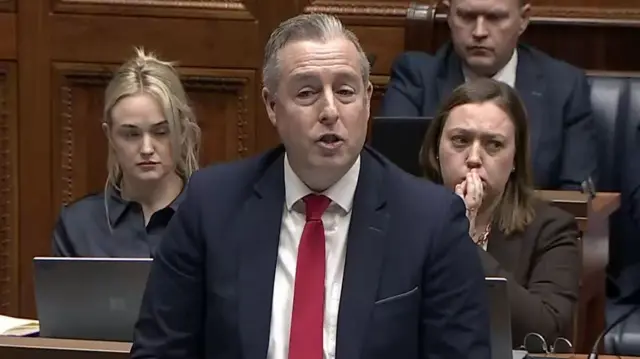 Paul Givan, dressed in suit and red tie addresses the assembly, with members seated behind him.