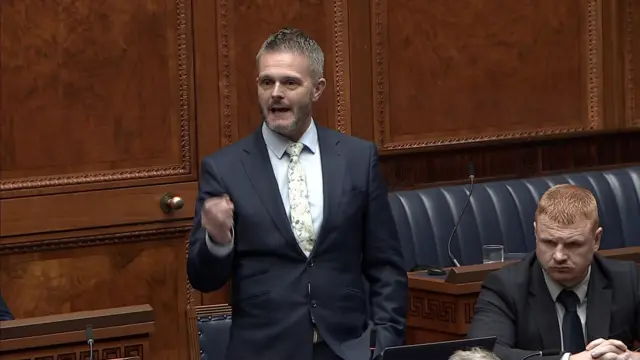 Robbie Butler, with arm outstretched, stands addressing the Assembly.